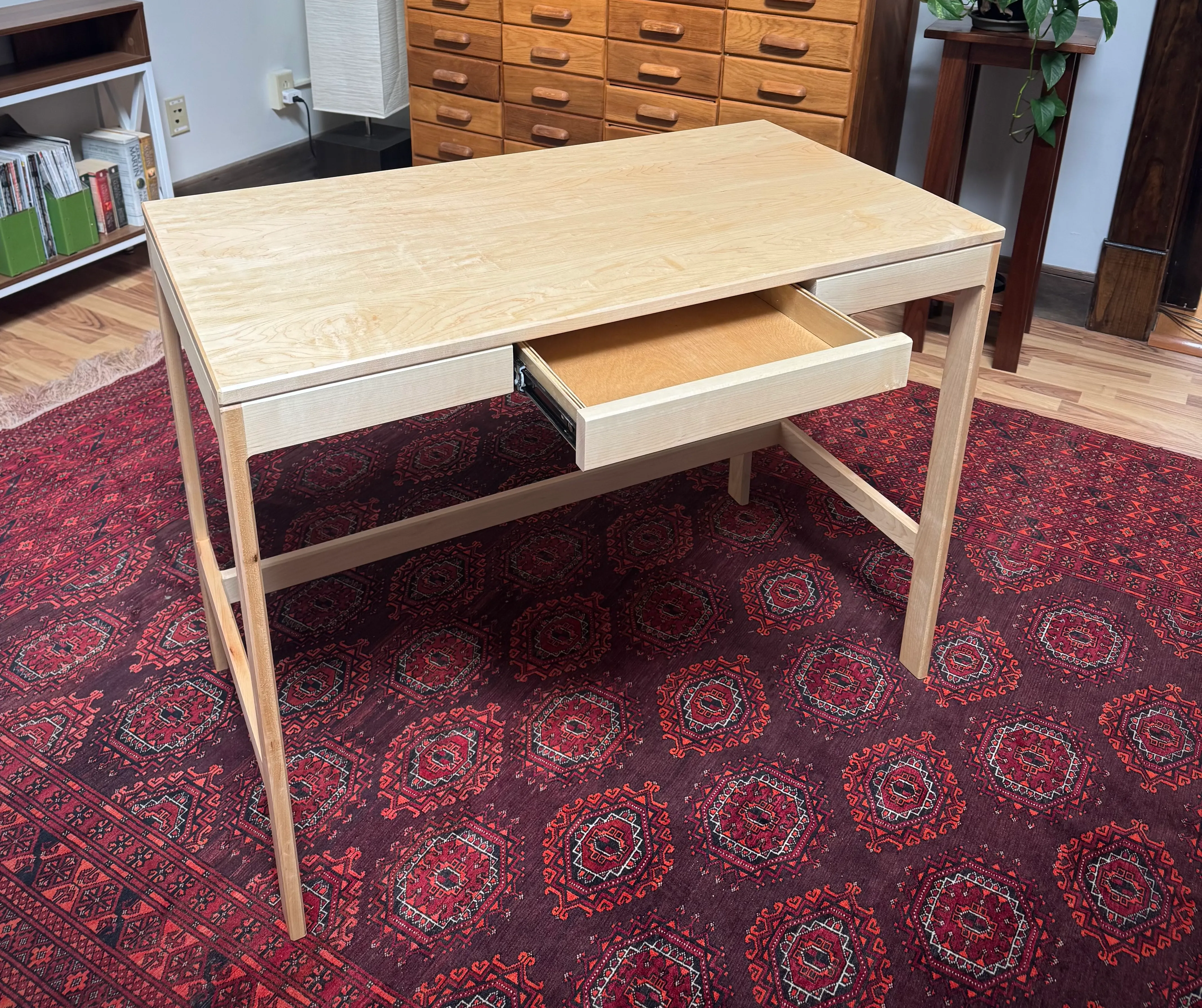 A solid maple writing desk with tapered legs and an open drawer, standing on a red patterned rug.